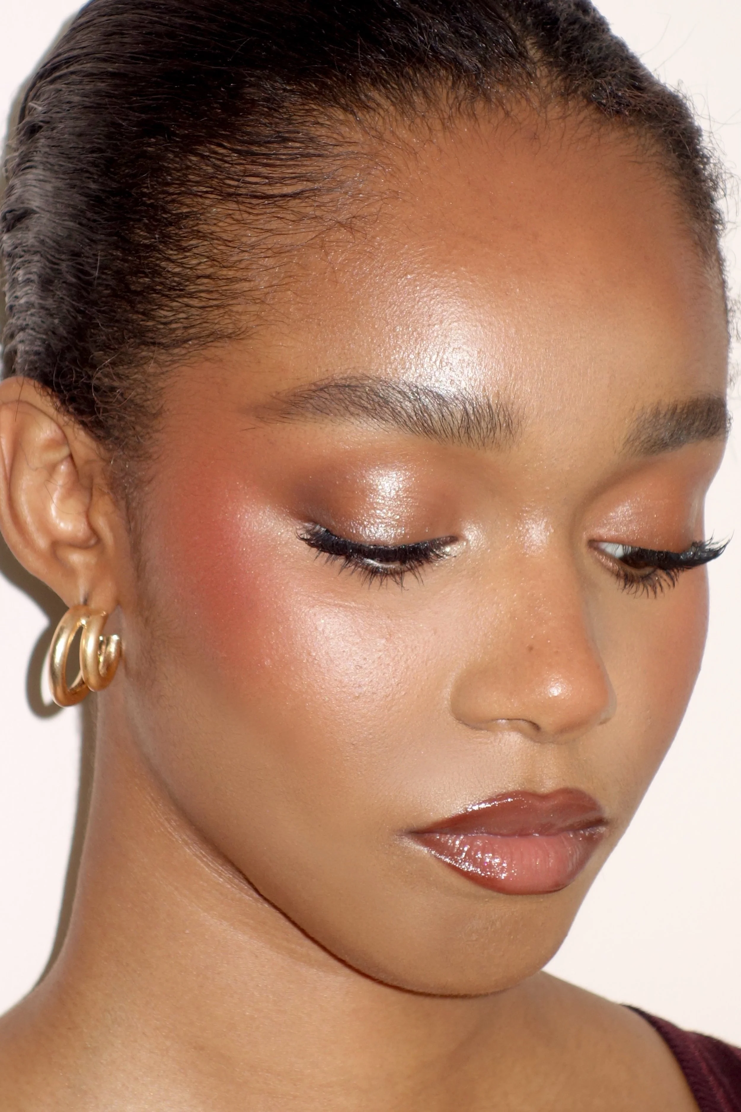Close-up of a woman's face focusing on her eyes, forehead, and cheek, with short dark hair, gold hoop earrings, and makeup including dark eyeliner, shimmering eyeshadow, and glossy lips.