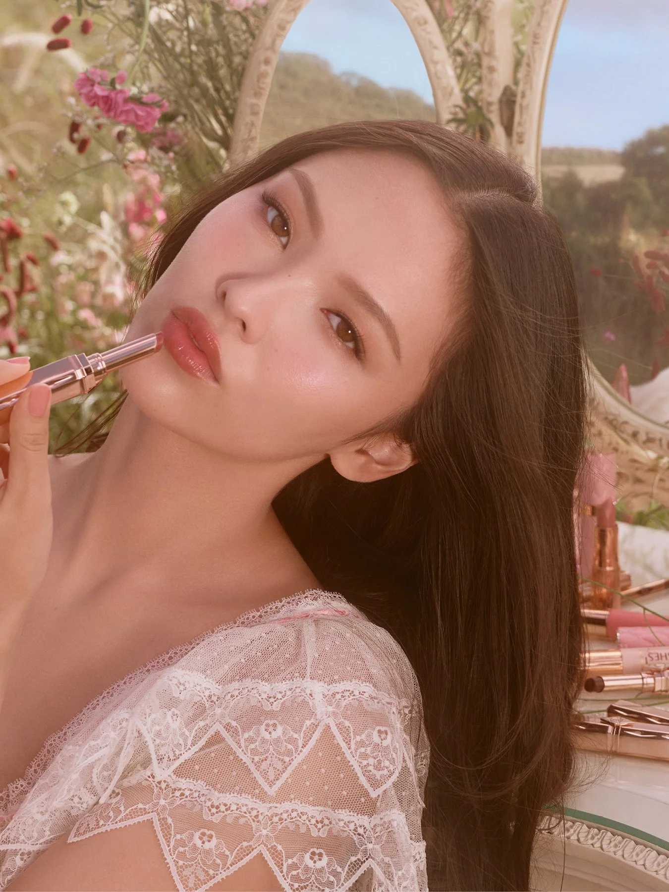 A woman with long brown hair applying lip gloss, sitting in a room with floral decor and makeup products in the background.