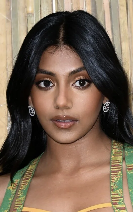 Close-up portrait of a woman with long black hair, wearing silver earrings, makeup, and a green traditional outfit with yellow accents, standing in front of bamboo background.