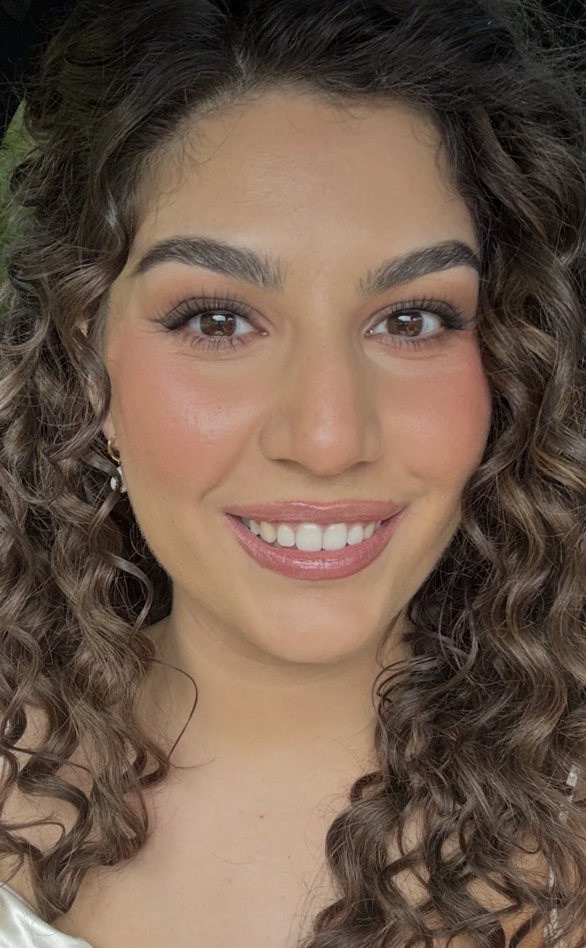 Close-up of a young woman with curly brown hair, brown eyes, and a bright smile, wearing natural makeup and small gold earrings.