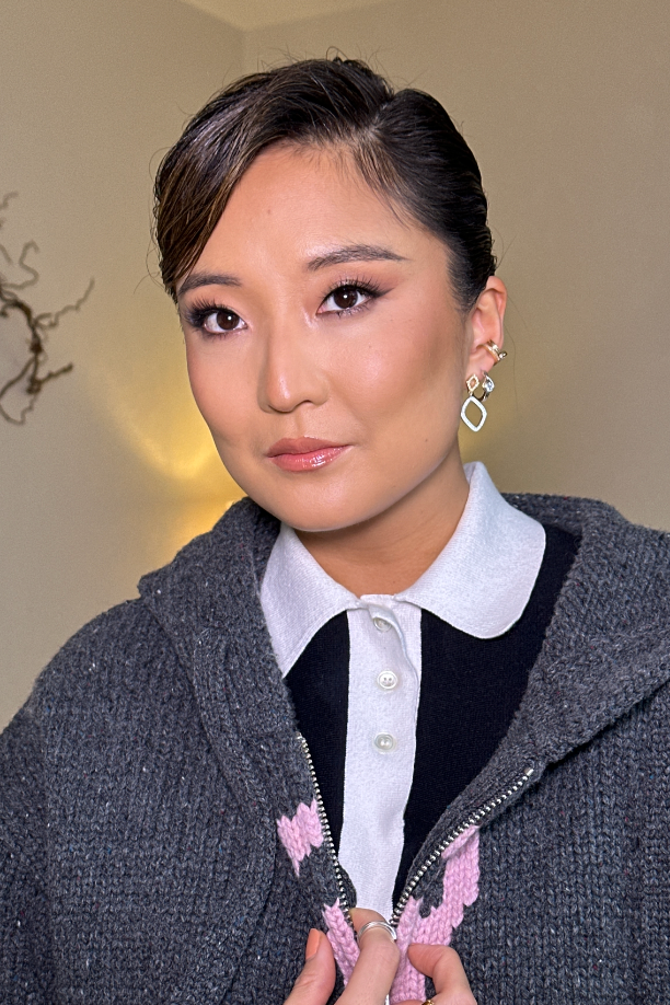 A woman with dark hair styled back, wearing a white collared shirt under a dark zip-up sweater, standing indoors with a blurred background, golden wall light, and a decorative branch.