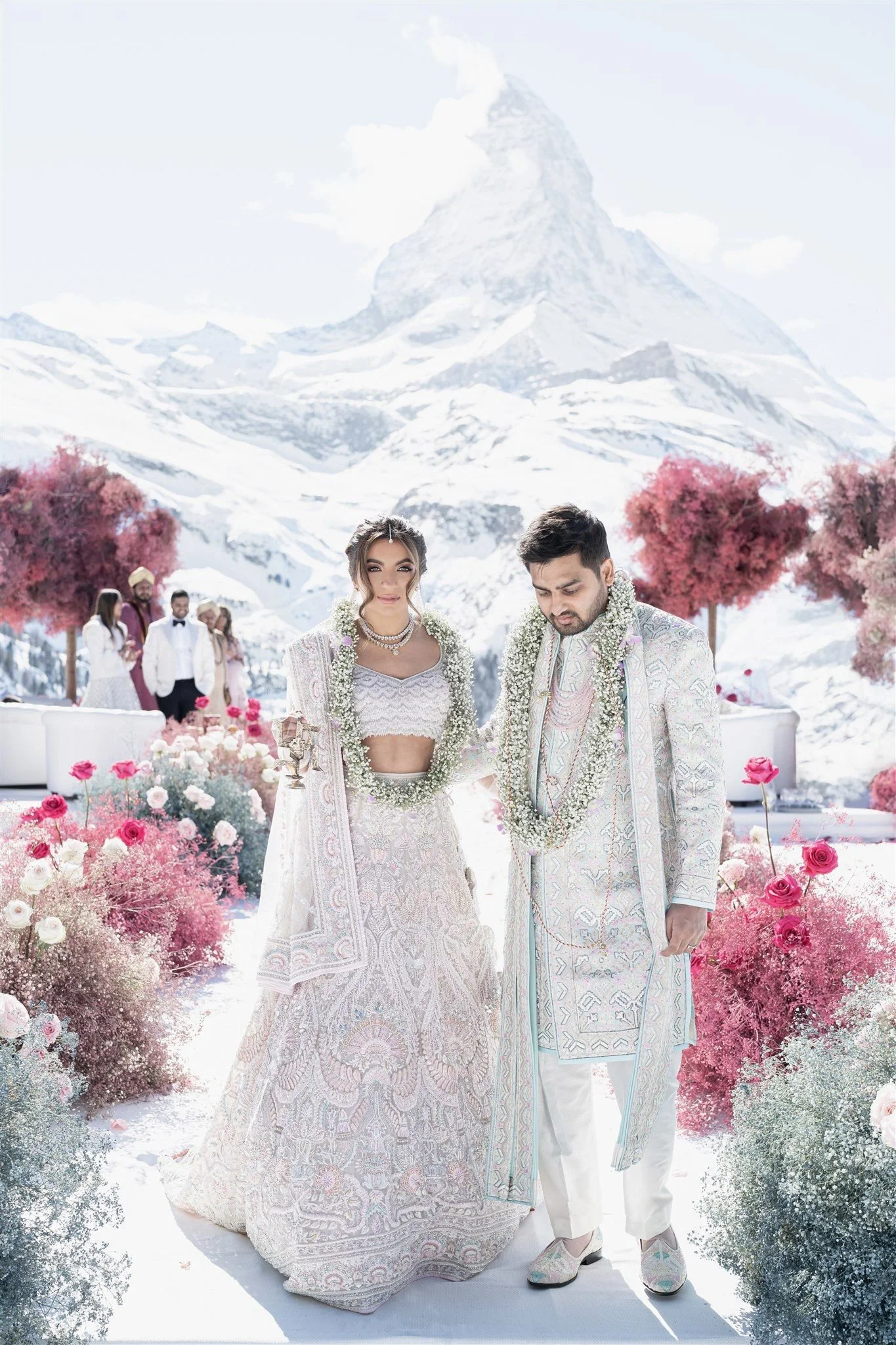 A wedding ceremony taking place outdoors in a snowy mountain landscape with pink and white floral arrangements. The bride and groom are dressed in traditional Indian wedding attire, standing closely with garlands around their necks. The bride is wear