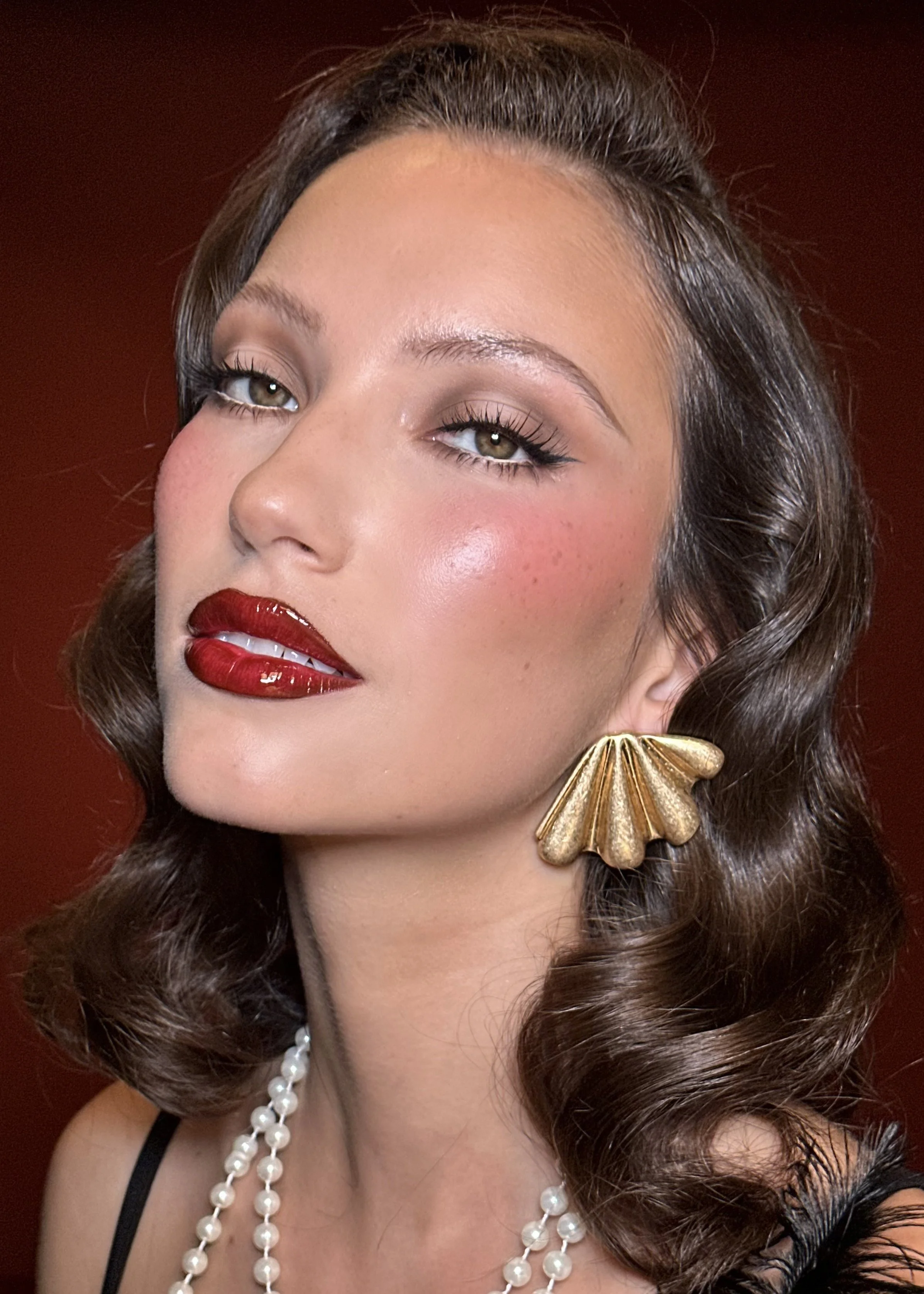 Close-up of a woman with vintage makeup, red lipstick, curled hair, gold shell earring, and pearl necklace, against a dark background.