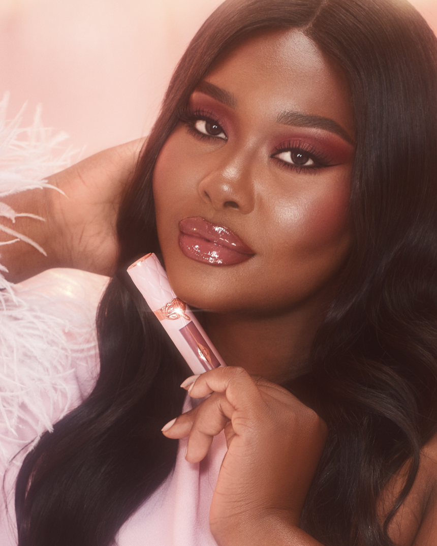 Close-up of a woman with long dark hair, glossy lips, and pink makeup, holding a pink lipstick, against a pink background with feathers.