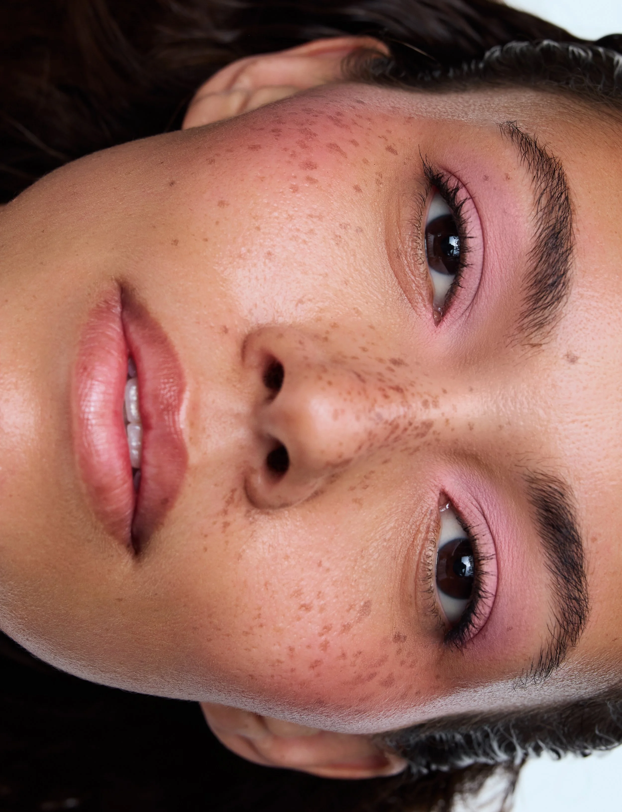 Close-up of a person's face lying down, showing detailed skin texture, freckles, dark brown eyes, and eyebrows.
