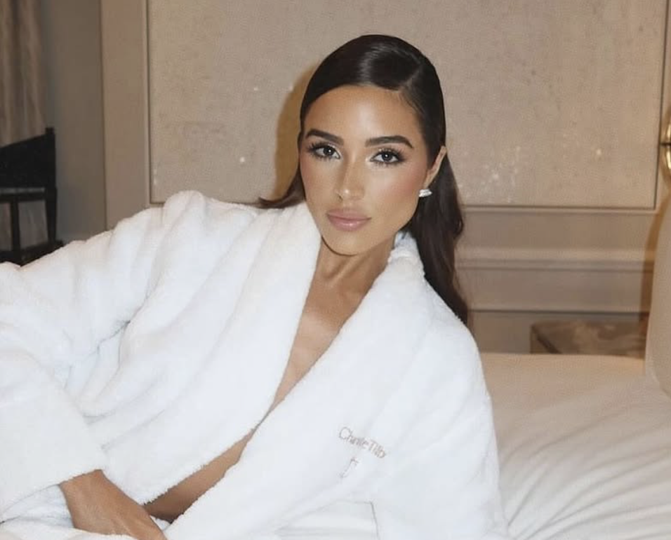 A woman with long dark hair, makeup, and earrings, wearing a white robe, sitting on a bed in a hotel room.