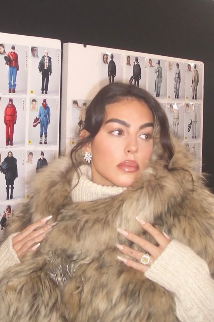 A woman with dark hair, wearing a cream turtleneck sweater, large floral earrings, rings, and a faux fur coat, posing in front of a display of fashion photos and sketches.