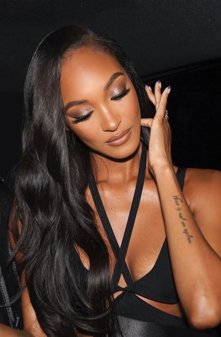 A woman with long black hair, closed eyes, and makeup, wearing a black outfit with thin straps, touching her face with her right hand.