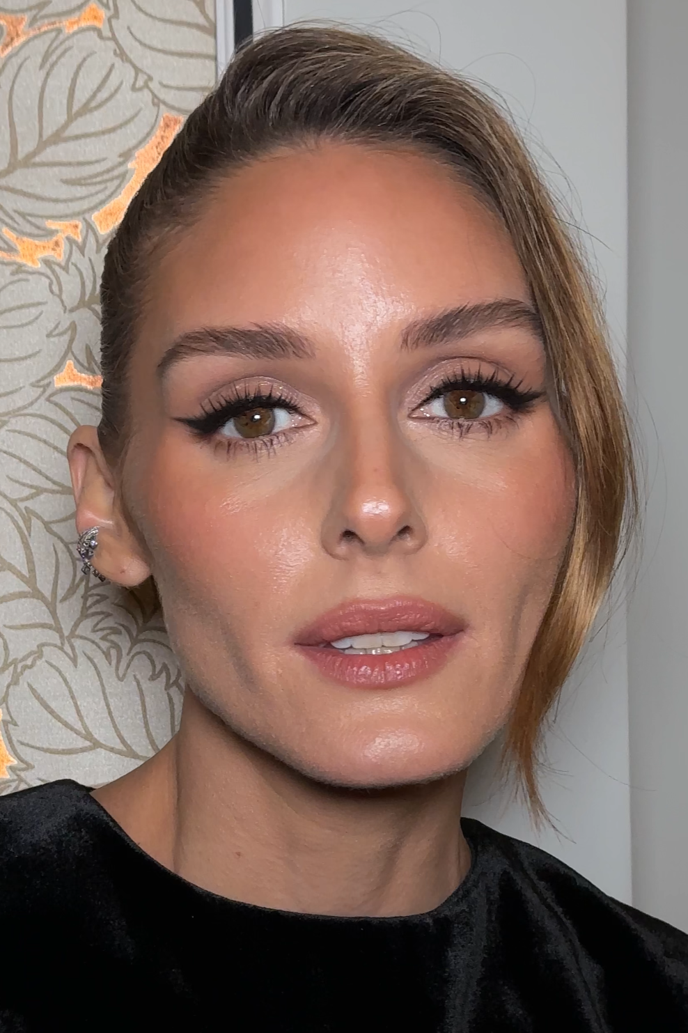 Close-up of a woman with styled hair, makeup, and earrings, standing in front of a patterned wall.