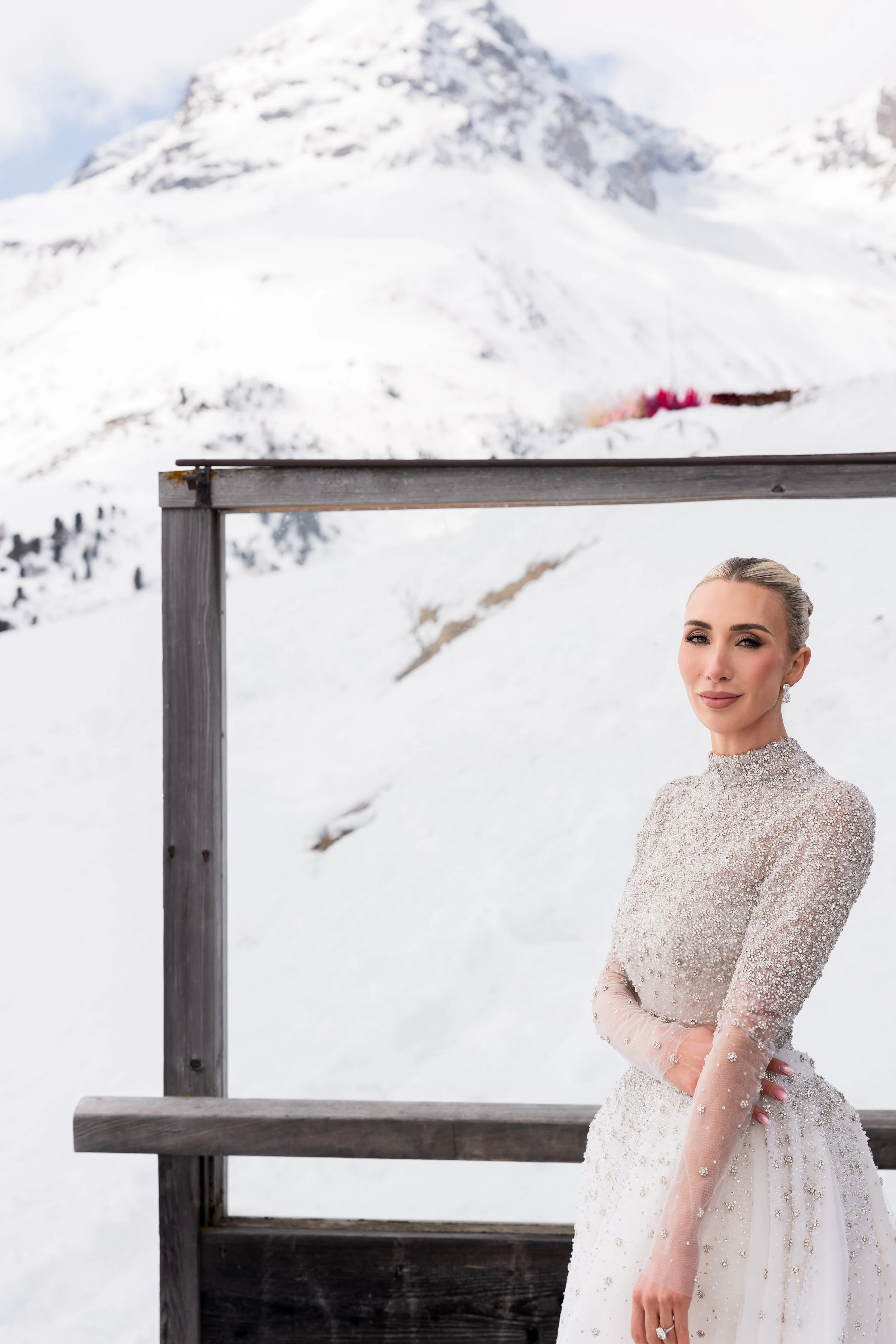 A woman in a sparkly wedding dress stands outdoors in a snowy mountain landscape, with part of a wooden frame in the foreground.