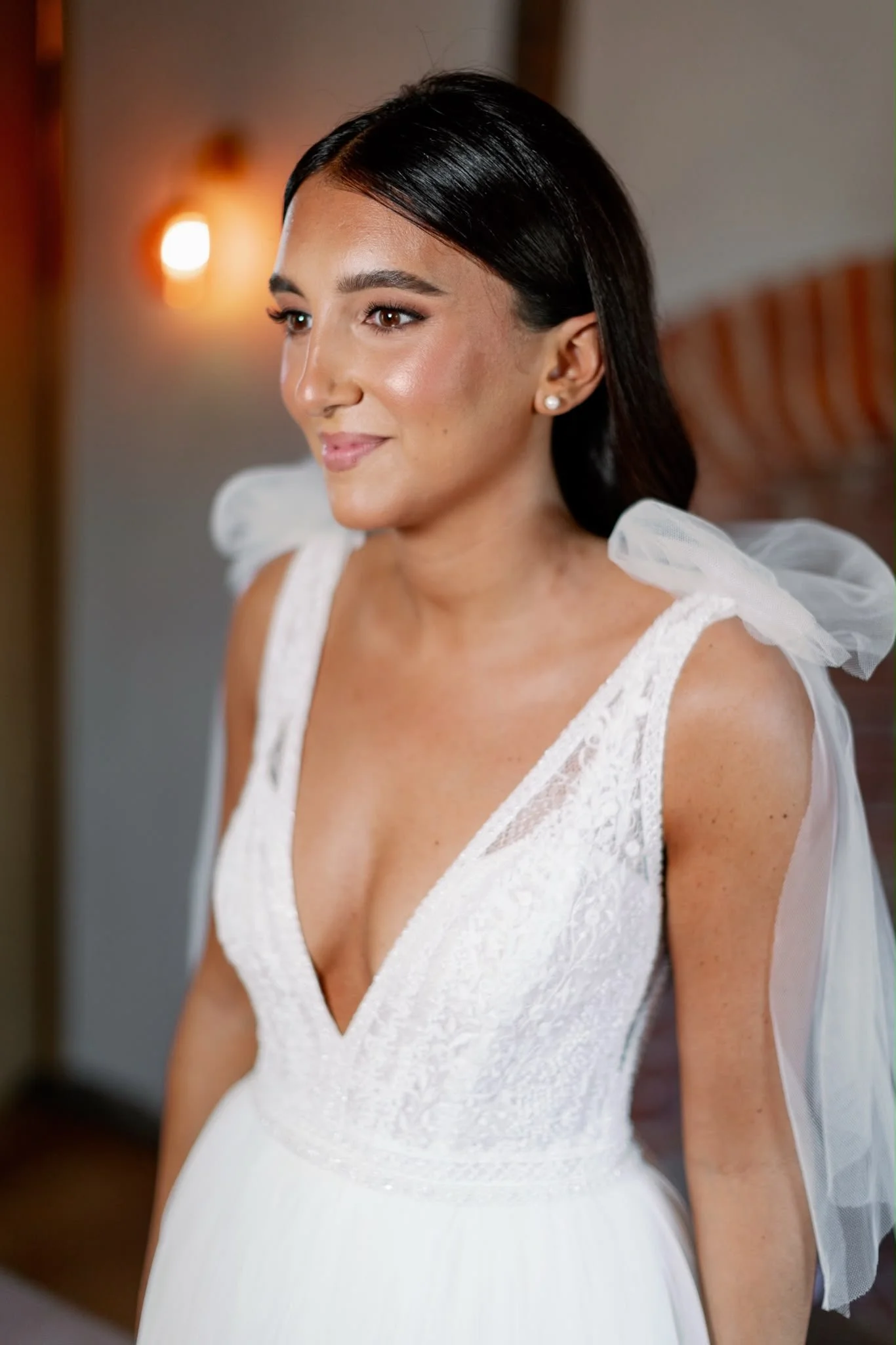 A woman with dark hair in a side profile, wearing a white dress with lace details and pearl earrings, smiling softly.