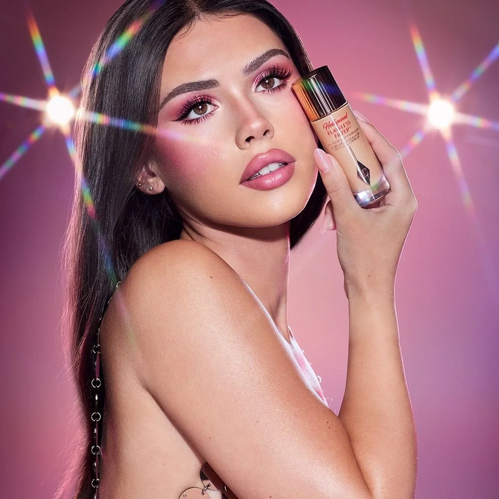 A woman with long dark hair holds a bottle of foundation near her face, with a pink background and rainbow light effects.