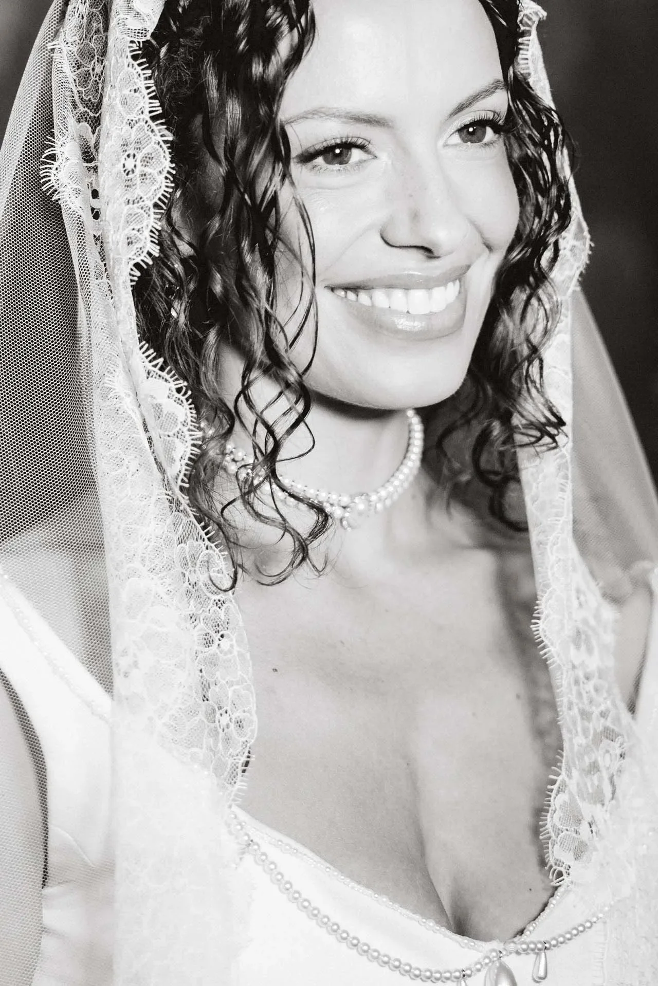 A black and white close-up portrait of a woman with curly hair, smiling, wearing a lace veil, a pearl necklace, and a pearl-edged dress, likely a bride.