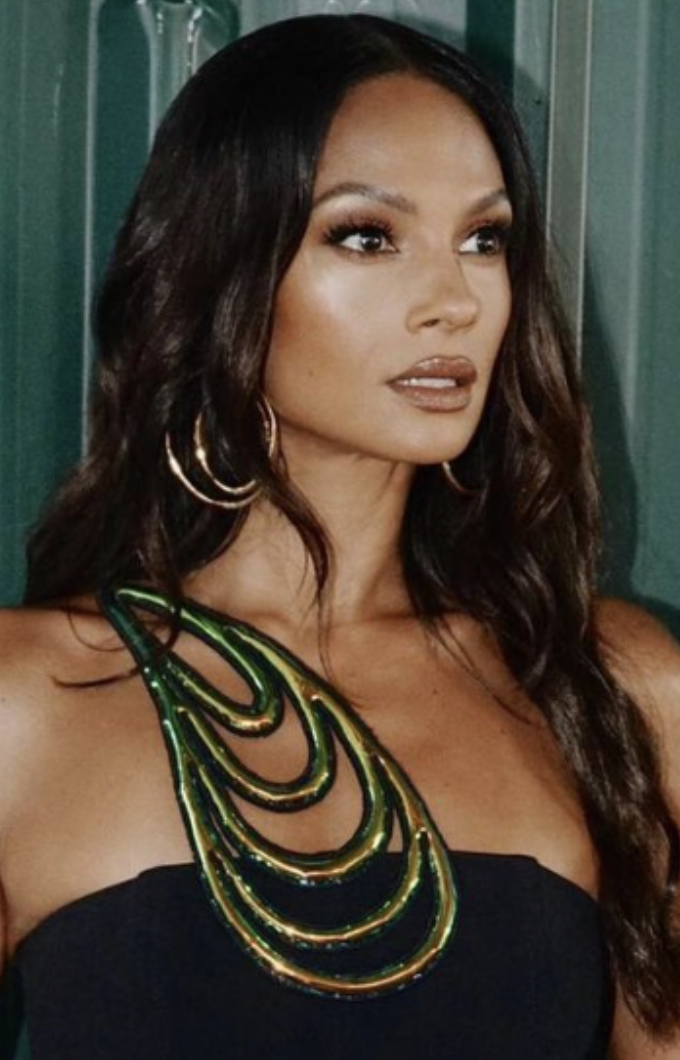 A woman with long dark hair, wearing large gold hoop earrings, a black strapless top, and a multi-layered green and gold necklace, standing against a green background.