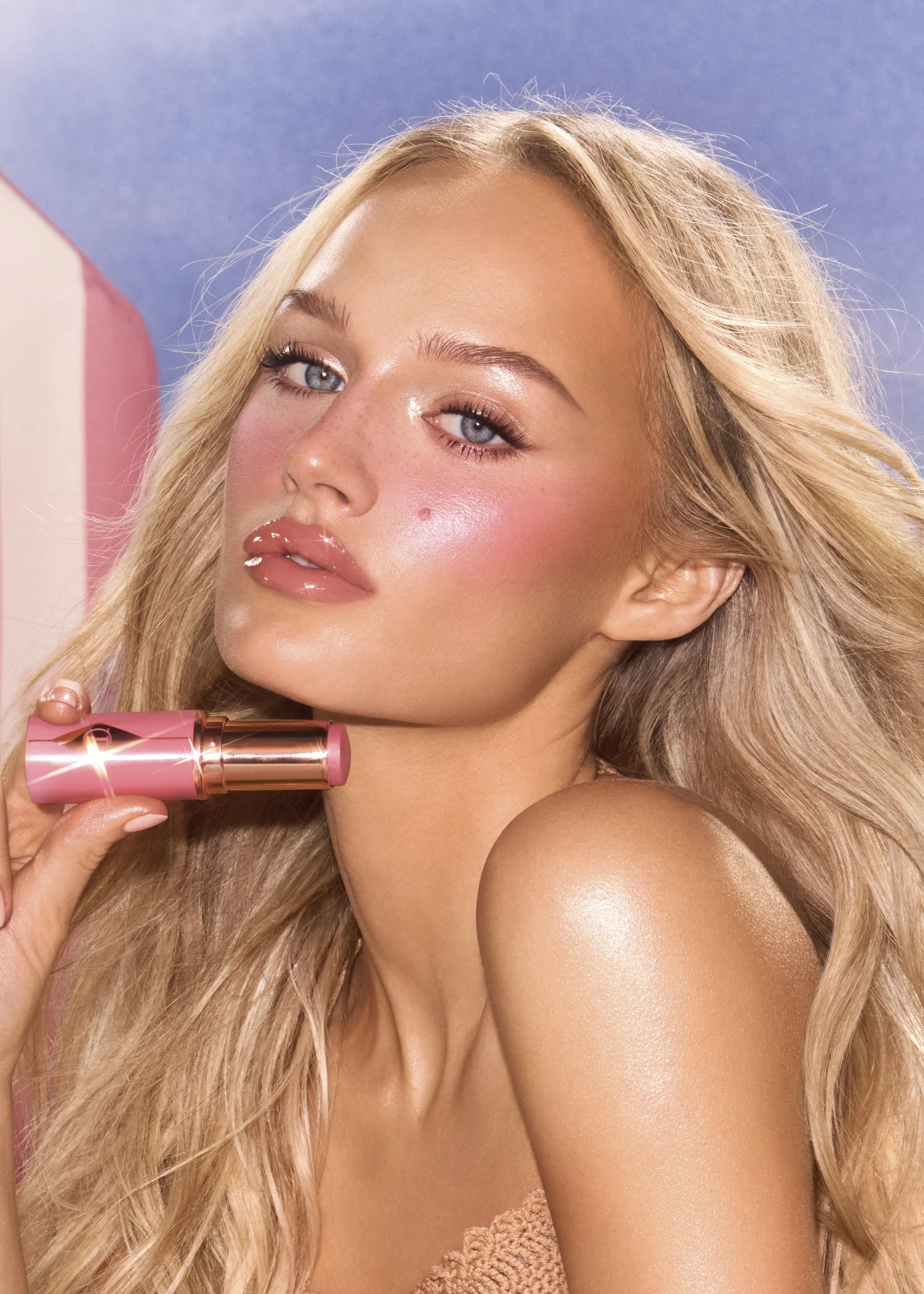 A woman with blonde hair and blue eyes holding a pink lipstick, with glossy lips and a soft pink blush on her cheeks, against a pastel background.