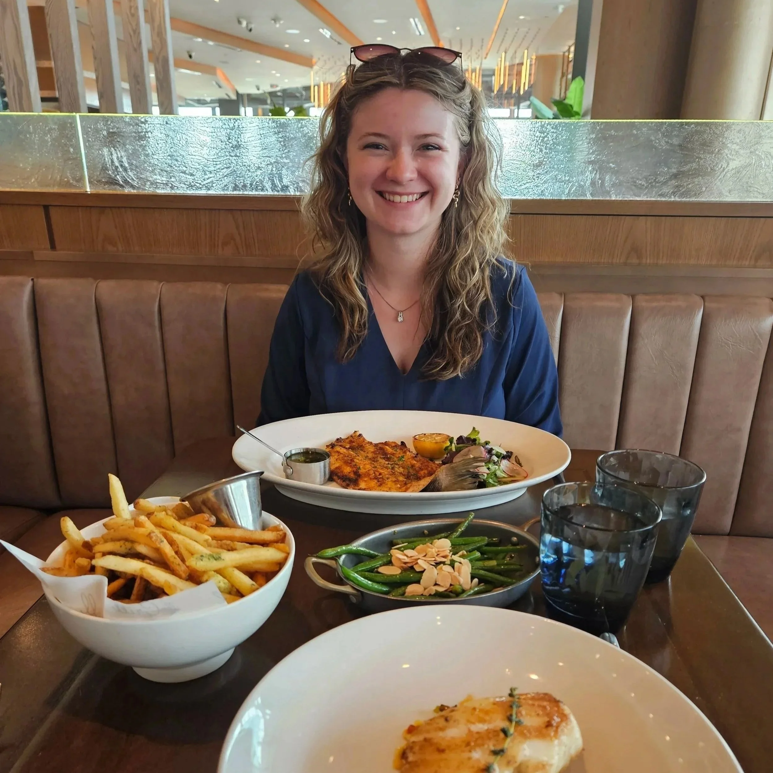 A woman sitting at a restaurant table smiling with sunglasses on her head. In front of her on the table are plates of French fries, a salad with sliced almonds, a grilled fish fillet, and a glass of water.