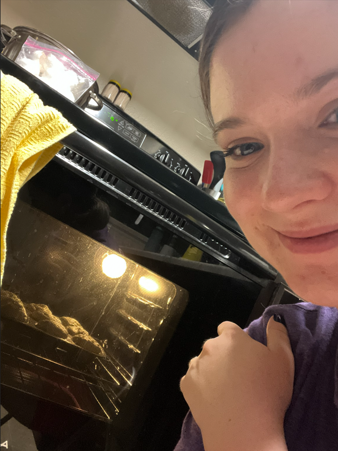 Person smiling near a countertop with a towel, a cooking appliance, and some containers, with an oven in the foreground showing baking cookies.