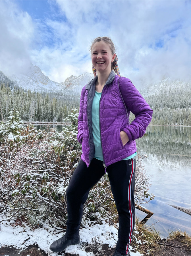A smiling young woman stands outdoors in front of a snowy mountain landscape with a lake, dressed in a purple jacket, black pants with red and white stripes, and hiking boots.