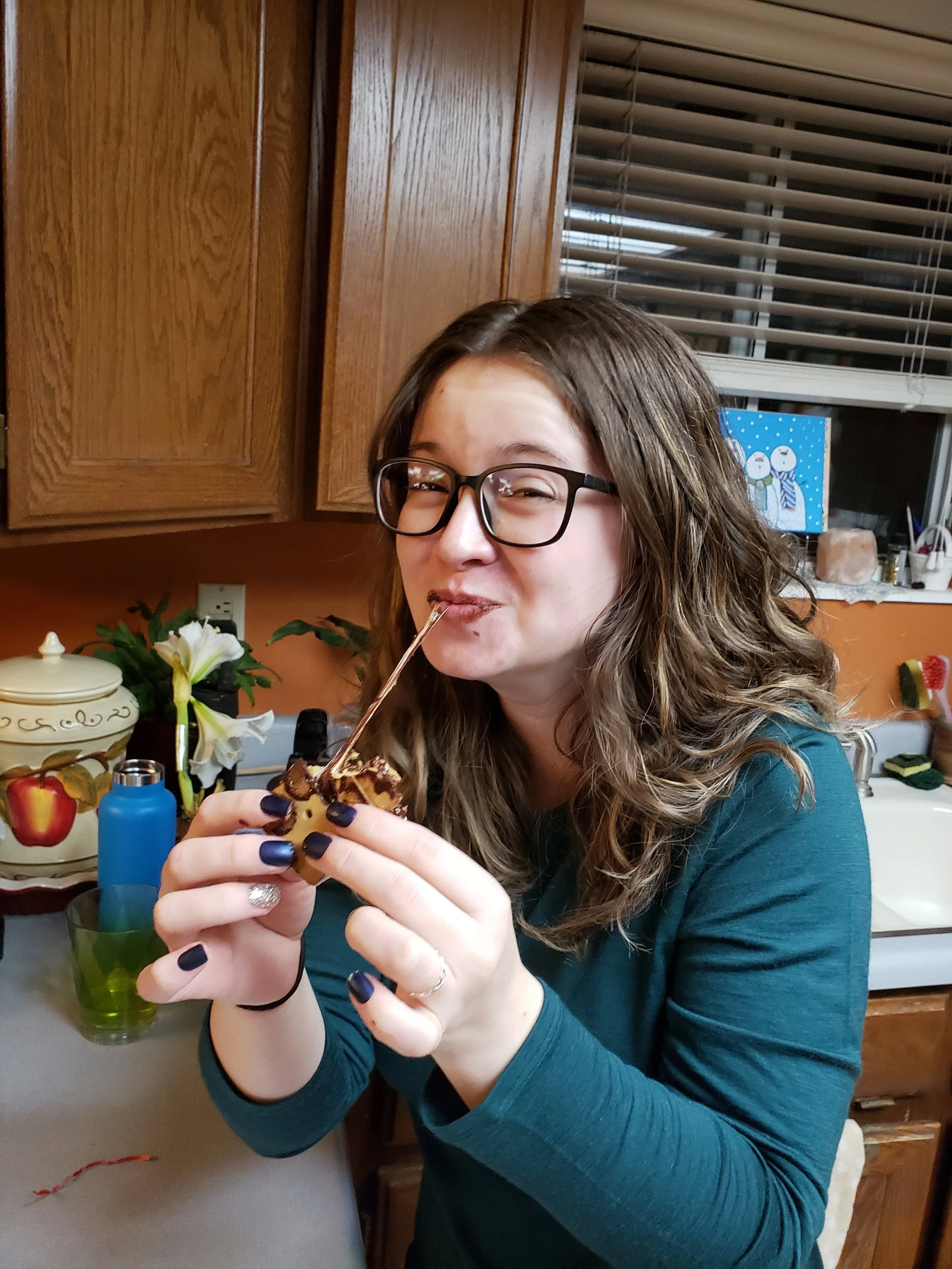 A young woman with glasses and long wavy brown hair is smiling and holding a dessert with chocolate topping. She is in a kitchen with brown wooden cabinets, a window with blinds, and decorative items on the counter.