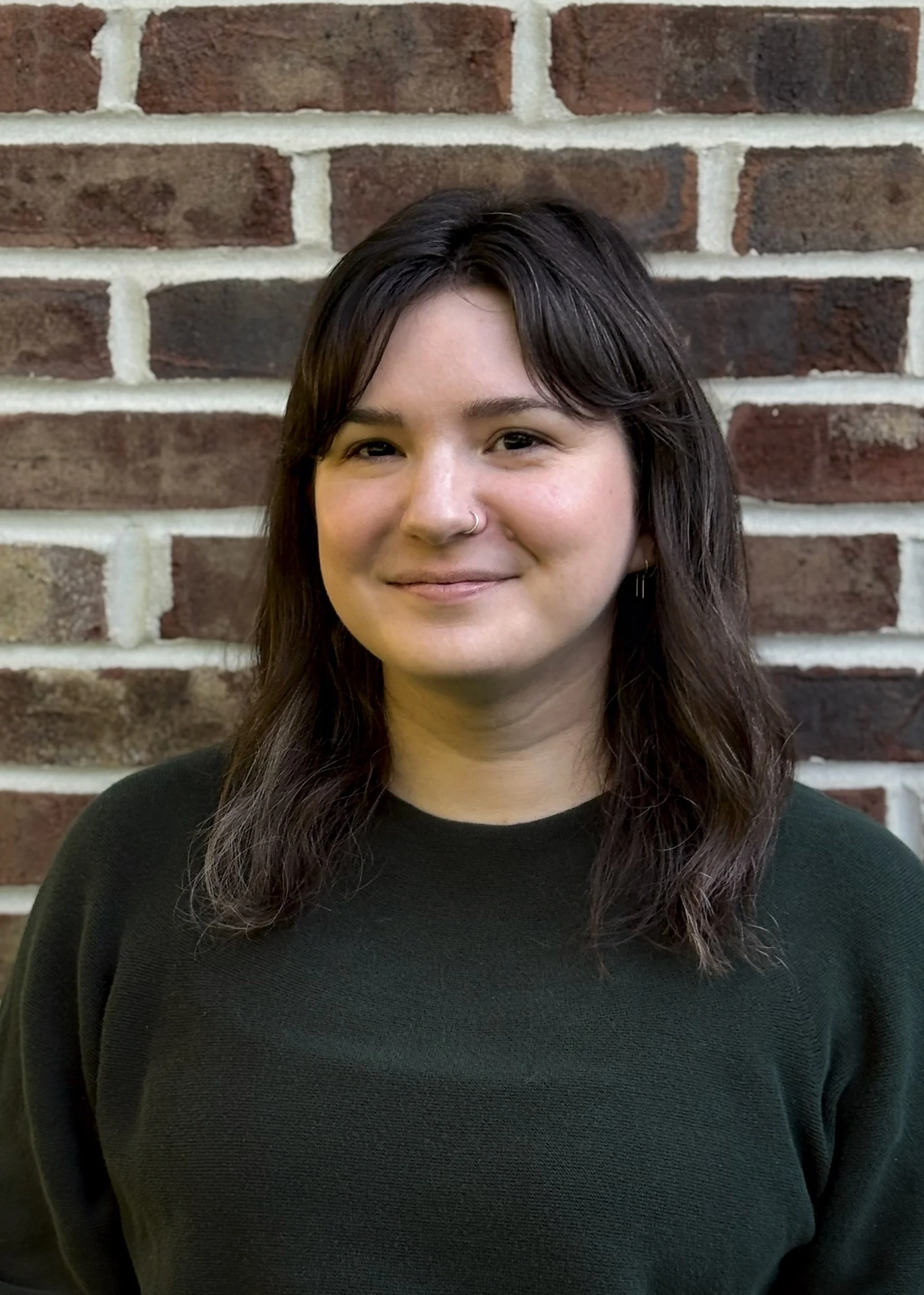 A person with dark hair, wearing a black sweater, standing against a brick wall background, smiling slightly.