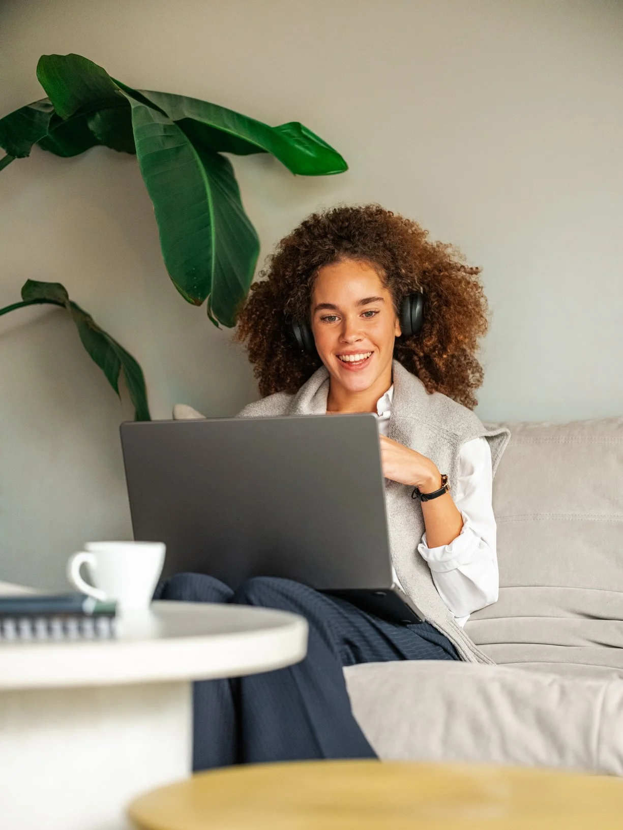 A woman with curly hair wearing headphones and a white shirt, sitting on a couch and smiling while looking at her laptop. There is a large green plant behind her and a table with a white cup in front.