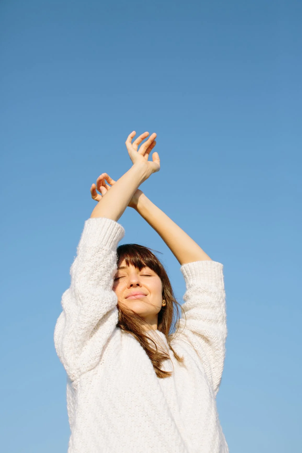 A woman with brown hair and closed eyes, smiling peacefully, wearing a white sweater, with hands raised and fingers crossed above her head, against a clear blue sky.