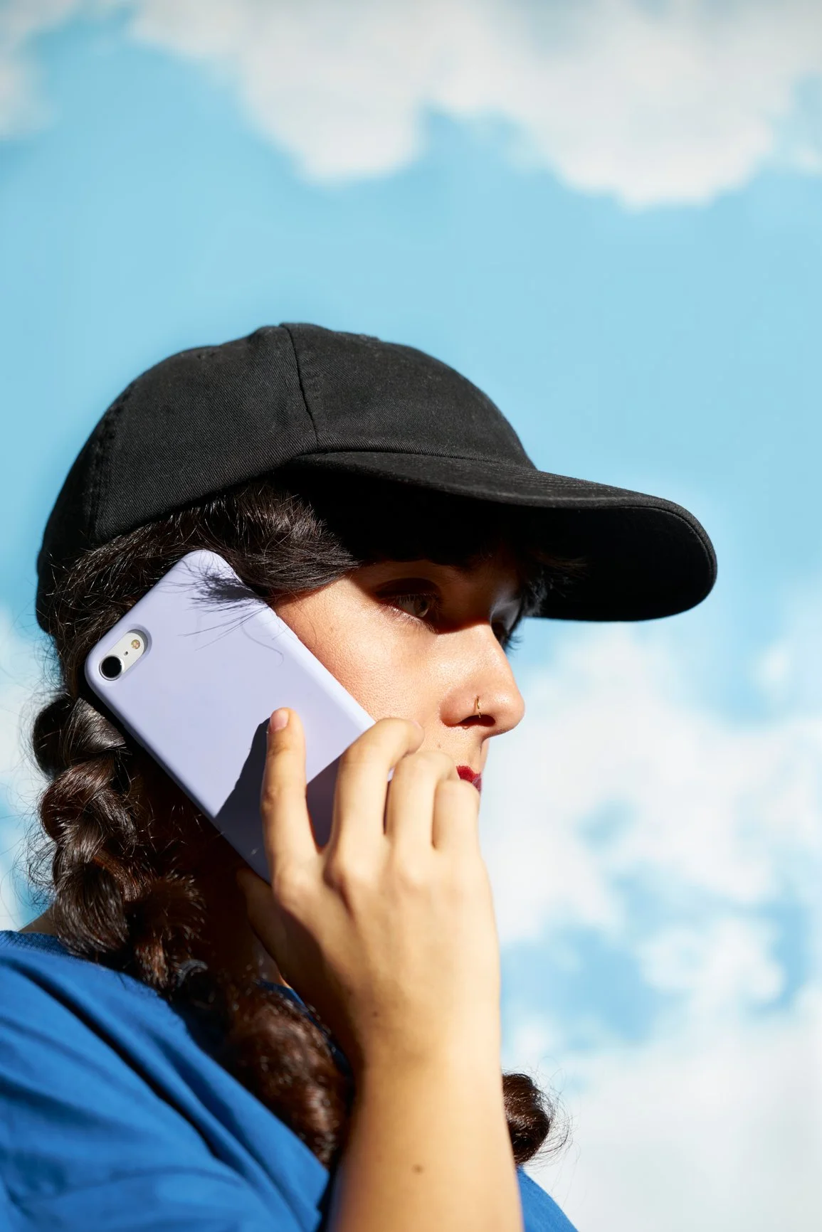 A woman wearing a black cap and blue jacket talking on a white phone against a blue sky with white clouds.