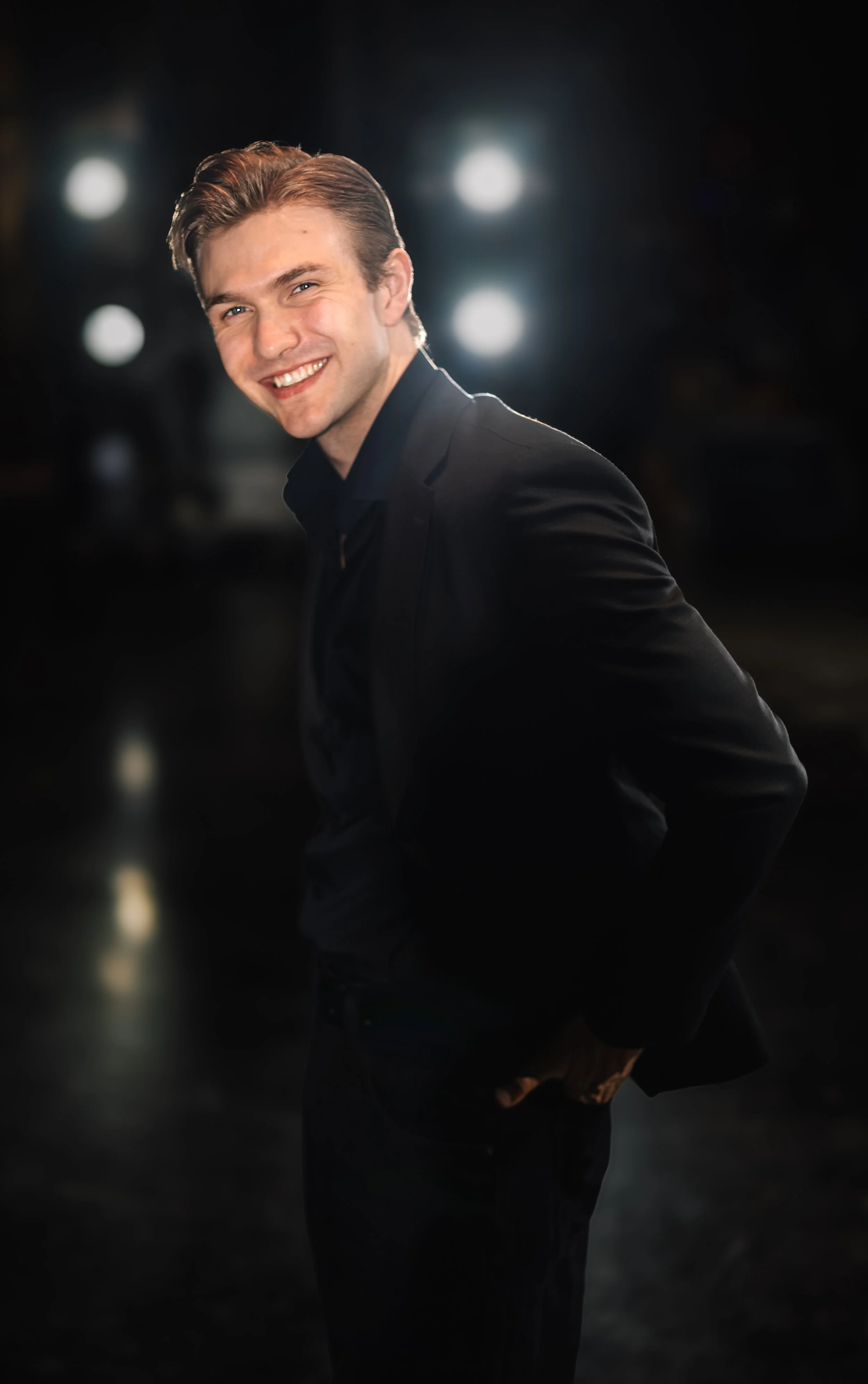 A young man with light brown hair styled back, smiling and looking at the camera. He is wearing a black suit jacket and shirt, standing in front of a dark background with blurry bright lights.