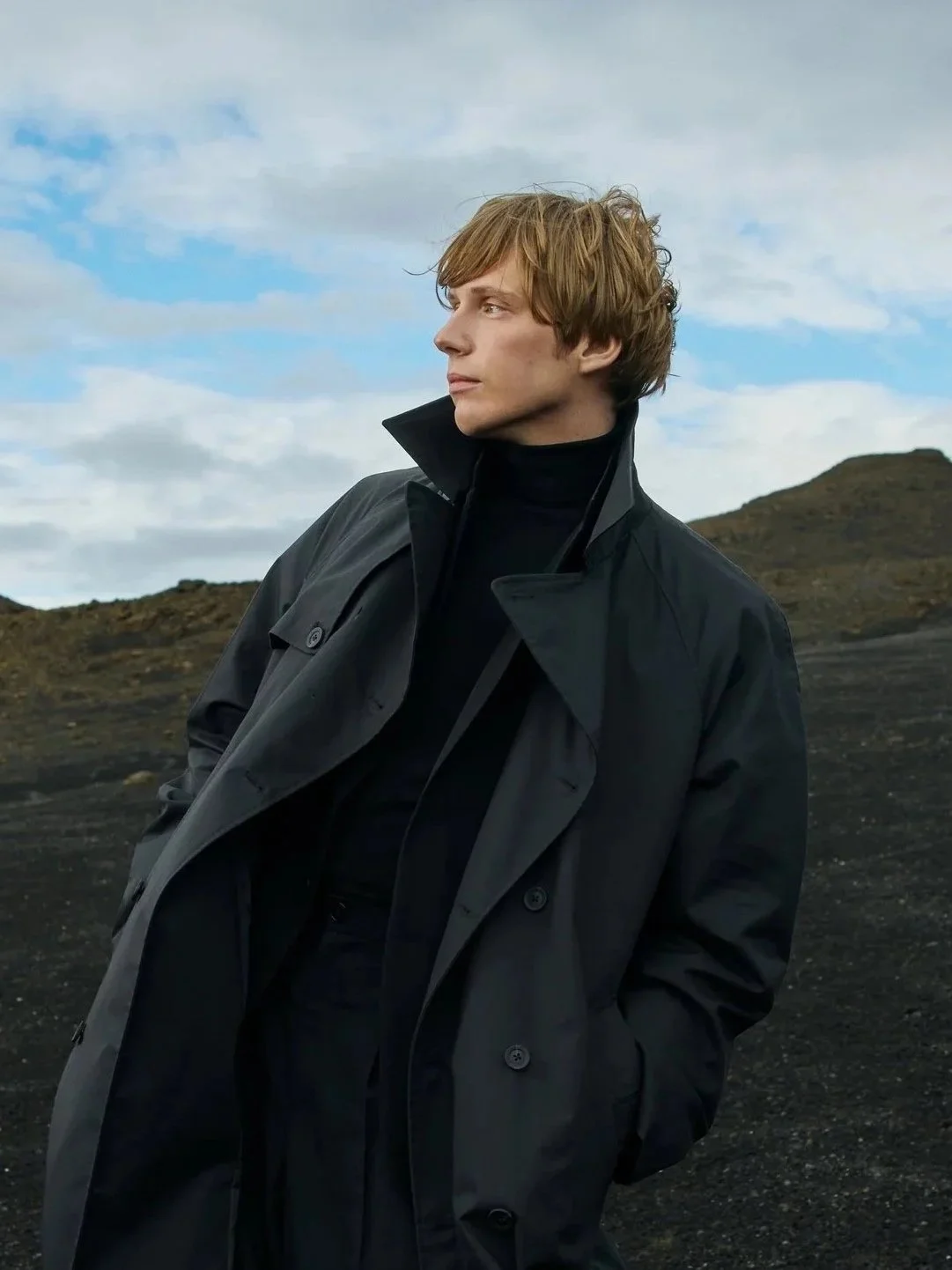 A young man with blonde hair wearing a black coat and turtleneck standing outdoors against a cloudy sky with rocky terrain in the background.