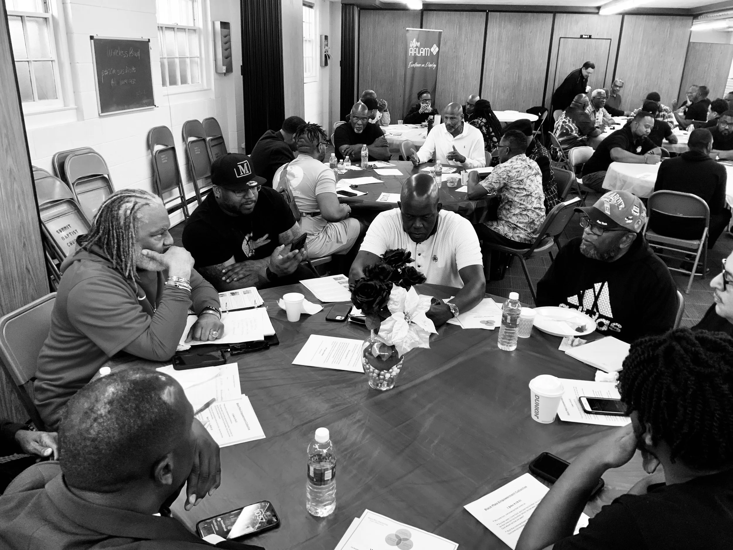 A black-and-white photo of a group of people seated around a large table engaging in discussion, with various documents, phones, and water bottles on the table, in a room with wood-paneled walls and a banner.