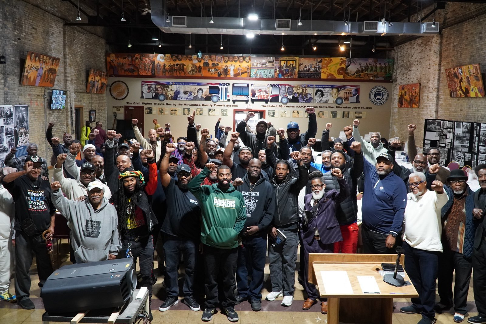 A large group of people gathered indoors in a historic museum or community center, raising their fists in a show of solidarity. The background features historical murals, photographs, and informational displays.