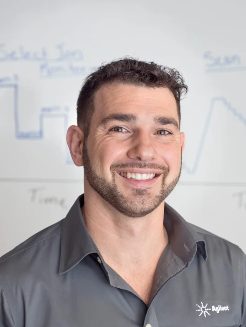 Smiling man with short curly hair and a beard wearing a dark gray shirt, standing in front of a whiteboard with handwritten notes and diagrams.