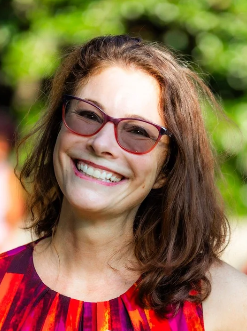Smiling woman with glasses and wavy hair outdoors, wearing a colorful top.