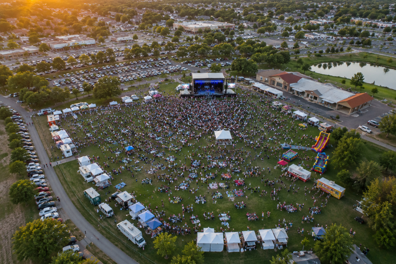 An outdoor concert event in a park with a large crowd gathered around a stage, surrounded by tents, food vendors, and inflatable attractions, with parking lots and trees in the background during sunset.