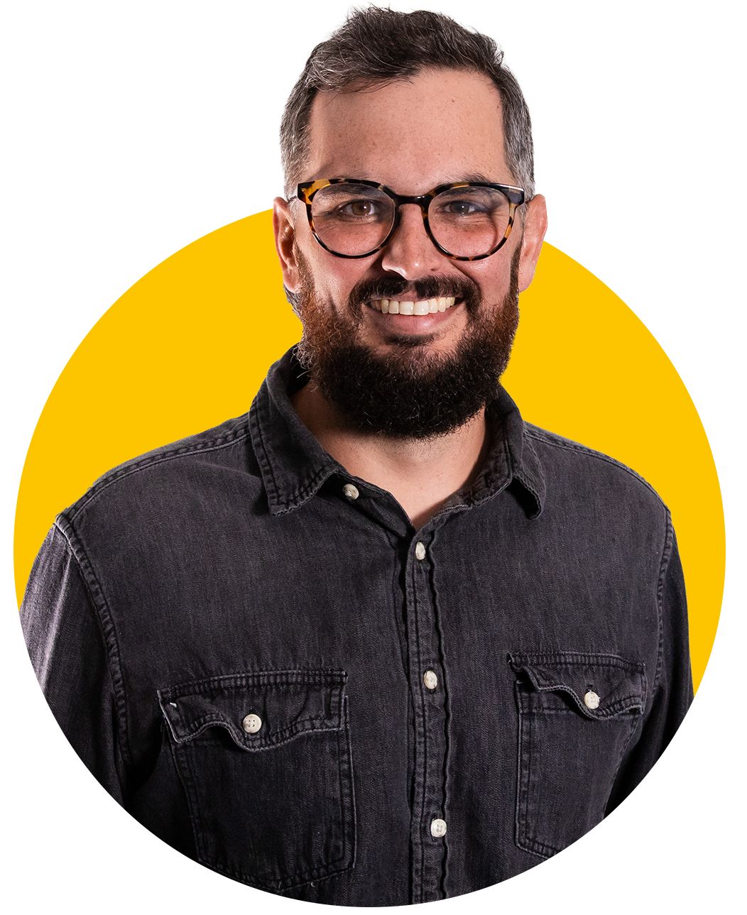 Portrait of a smiling man with glasses, a beard, and a dark denim shirt, standing against a yellow background.