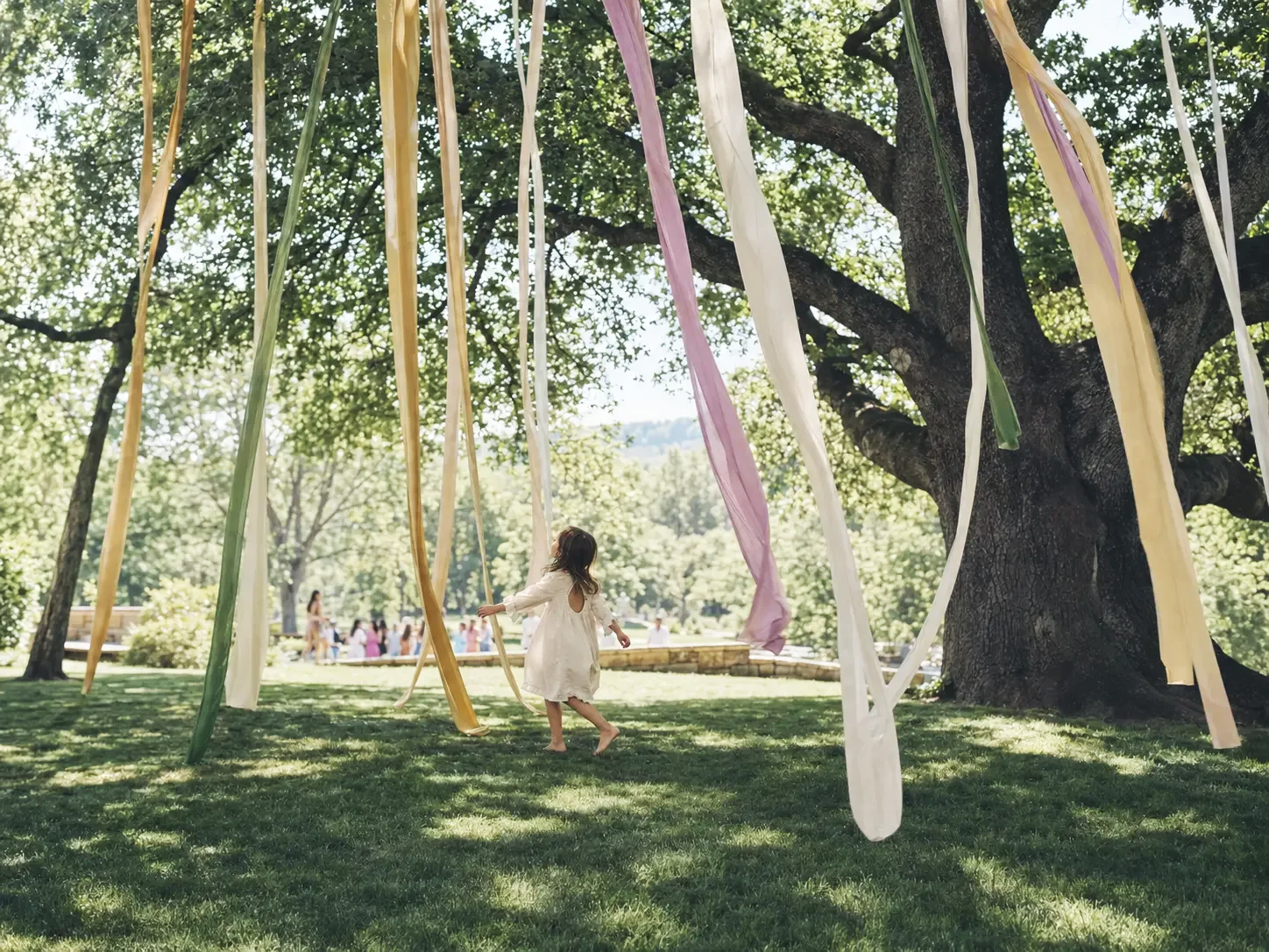 Young girl playing beneath a tree decorated with colorful ribbons at an outdoor gathering.