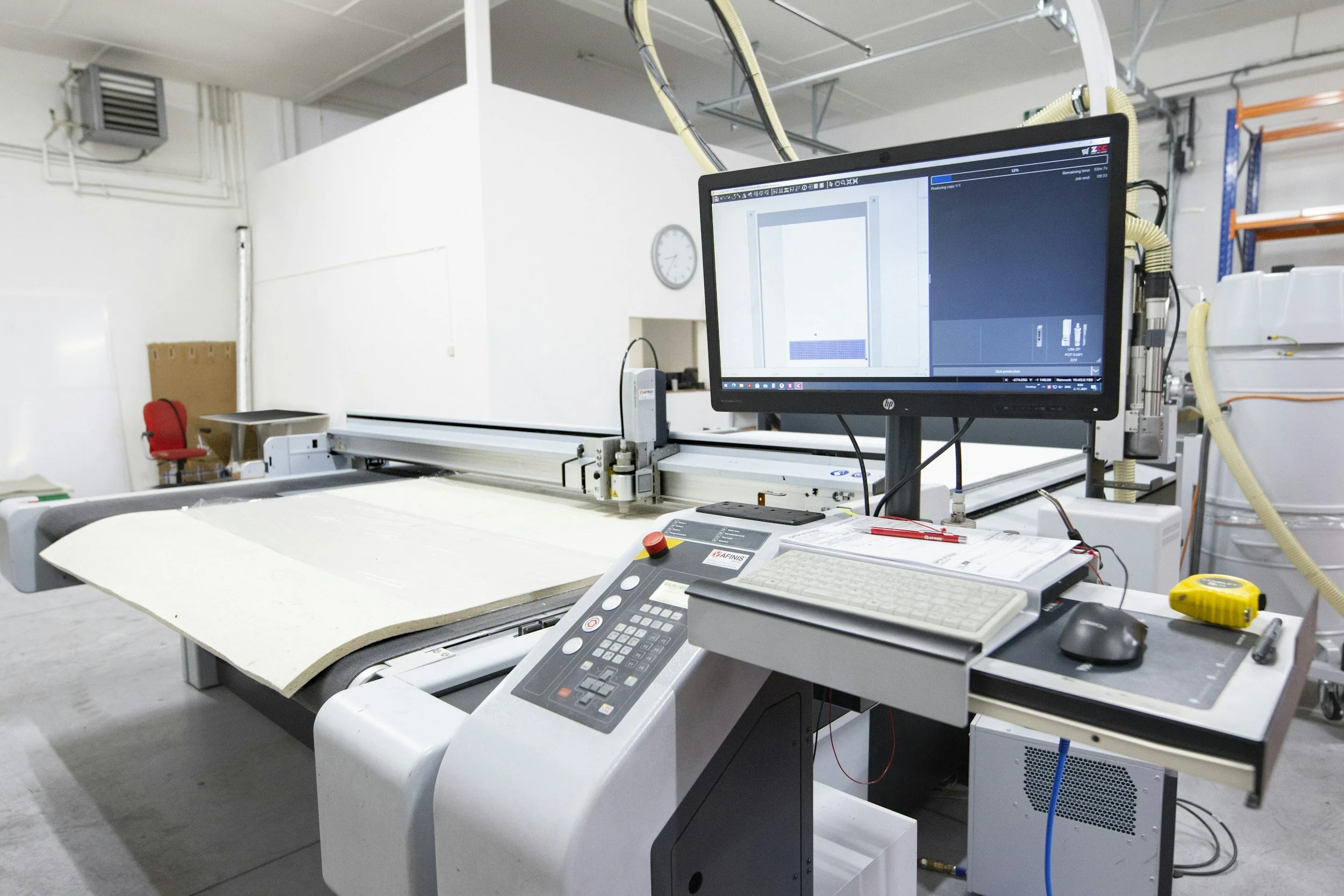 Industrial workspace with computer monitor displaying design software, control panel, yellow and black tools, and equipment on a white table in a clean, well-lit room with white walls and a clock.