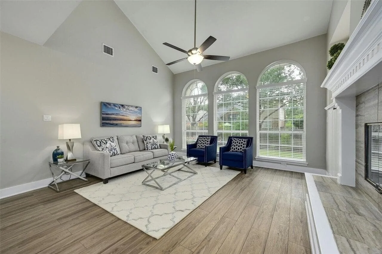 Living room with three large arched windows, a gray sofa, two blue armchairs with patterned pillows, a glass coffee table, two side tables with lamps, a painting above the sofa, a decorative rug, ceiling fan, and a fireplace with white molding.