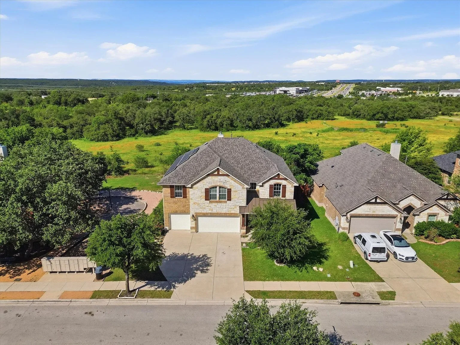 Aerial view of two suburban houses with driveways, surrounded by green lawns and trees, with open fields and a highway in the background under a partly cloudy sky.