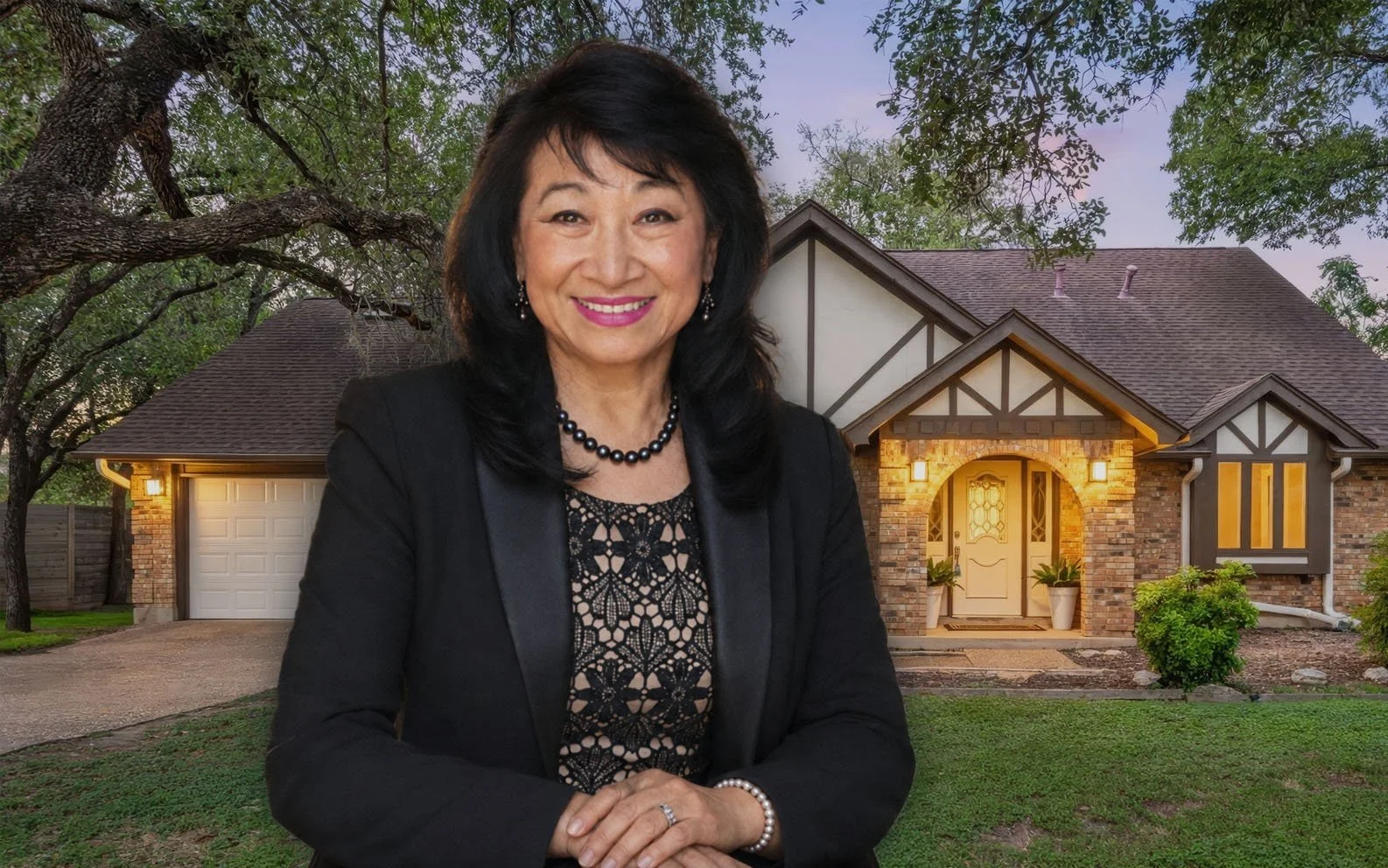 A woman with black hair smiling, dressed in a black blazer and lace top, standing in front of a brick house with a well-maintained lawn and trees in the background.