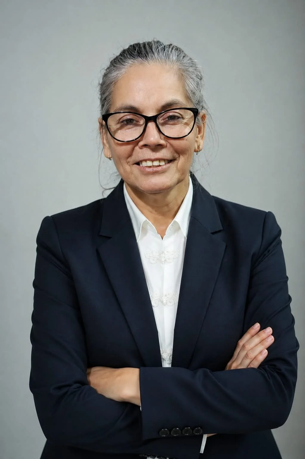 Mujer de cabello gris recogido, gafas negras, arreglada con traje negro y blusa blanca, sonriendo con brazos cruzados, en un fondo gris.
