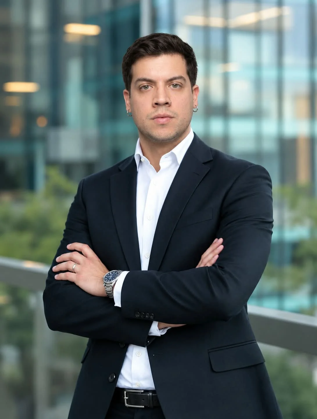 Hombre joven y bien vestido con chaqueta negra, camisa blanca y reloj, de cabello oscuro, de aspecto serio, con brazos cruzados frente a un fondo de oficinas con ventanas de vidrio.