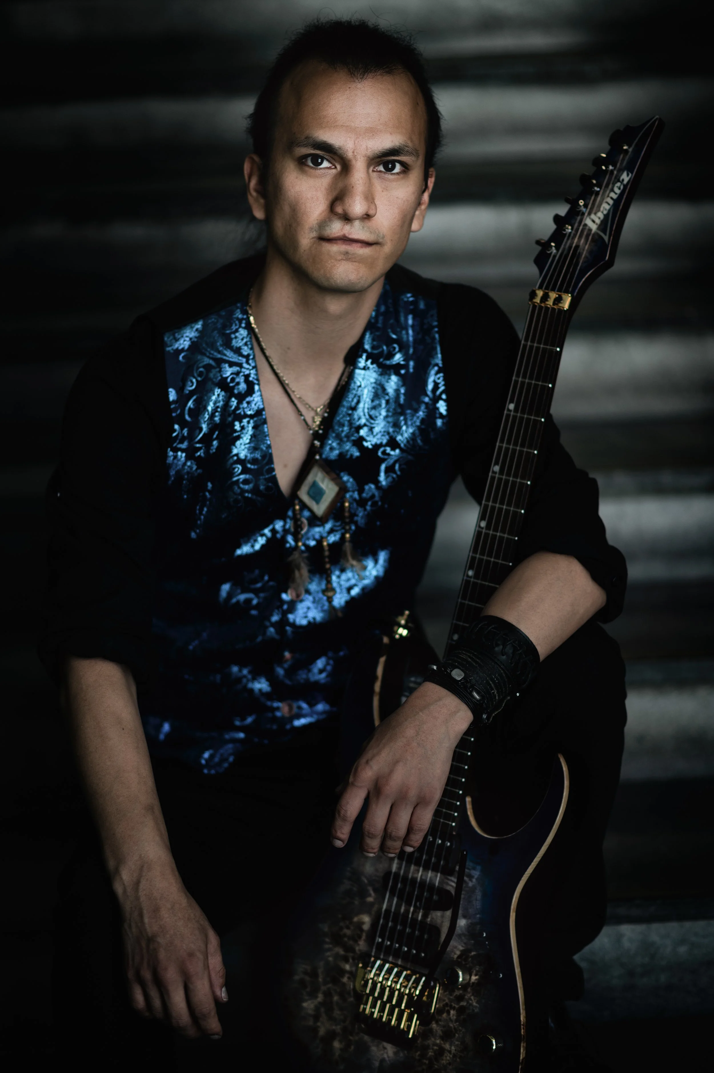 -JAIME- picture in Above and Beyond signature outfit of blue shiny west, universe necklace pendant, rock pants and signature wristbands. Holding his universe guitar with gold hardware, in front of a dark, textured background.
