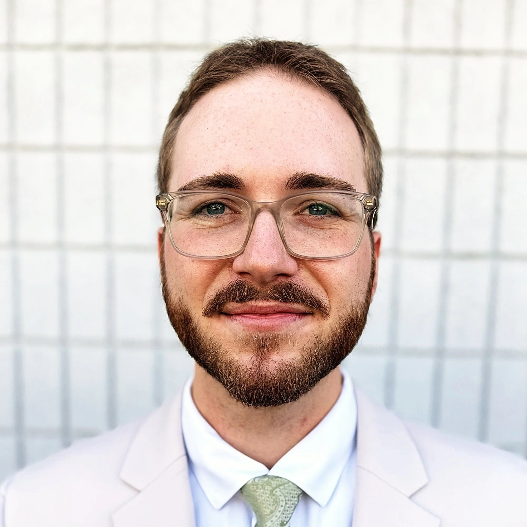 Close-up of a smiling man with glasses, wearing a white dress shirt and a paisley tie, with a light-colored brick wall background.