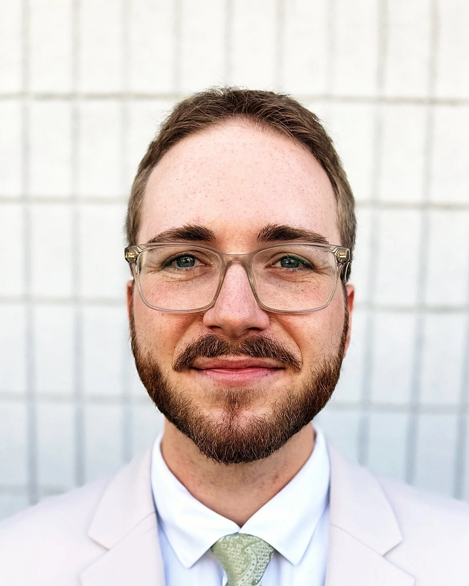 Close-up portrait of a man with short brown hair, a beard, and glasses, dressed in a white suit and green tie, smiling against a grid-patterned wall background.
