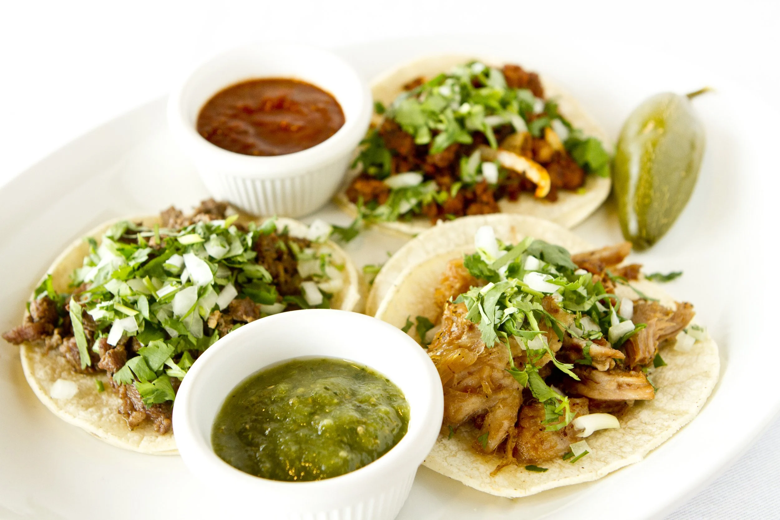 Close-up of three tacos filled with seasoned meat, topped with chopped onions and cilantro, served with green salsa and brown sauce in small white cups on a white plate.