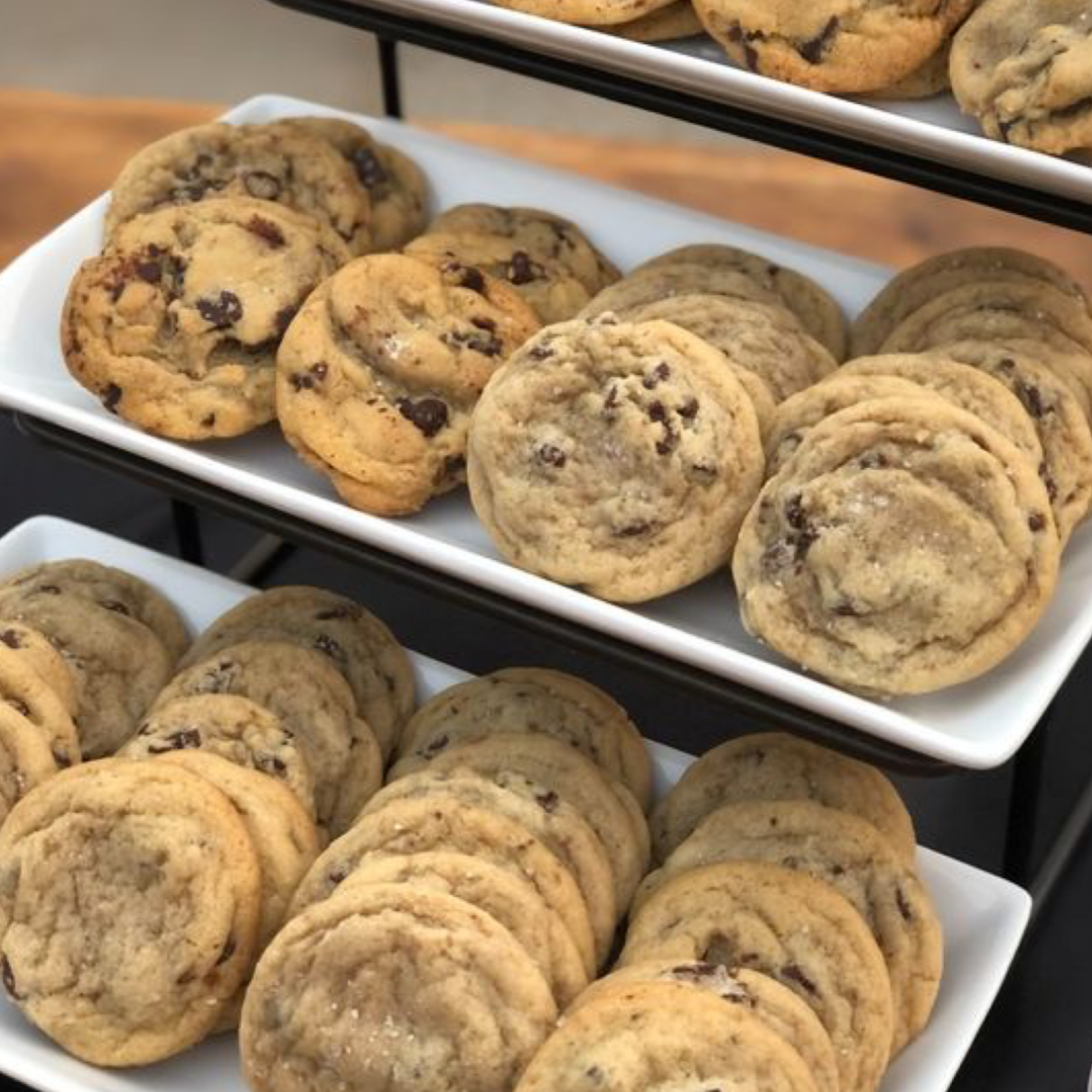 Three-tier white dessert display filled with homemade chocolate chip cookies. The cookies are golden brown with visible chocolate chunks and chips, arranged neatly on rectangular platters for an inviting bakery-style presentation.