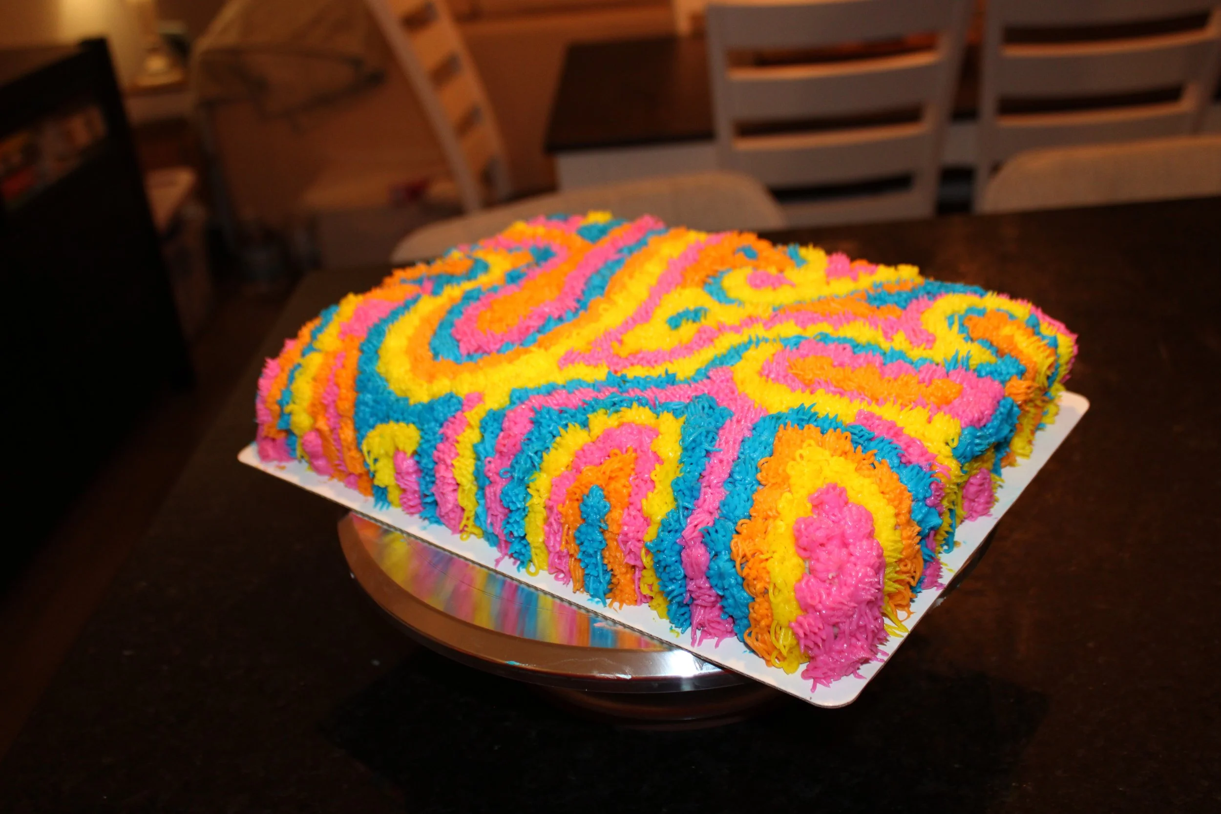Colorful shag cake decorated with bright neon pink, blue, yellow, and orange buttercream piped in a textured swirl pattern. The square cake sits on a white cake board and turntable on a dark kitchen countertop.