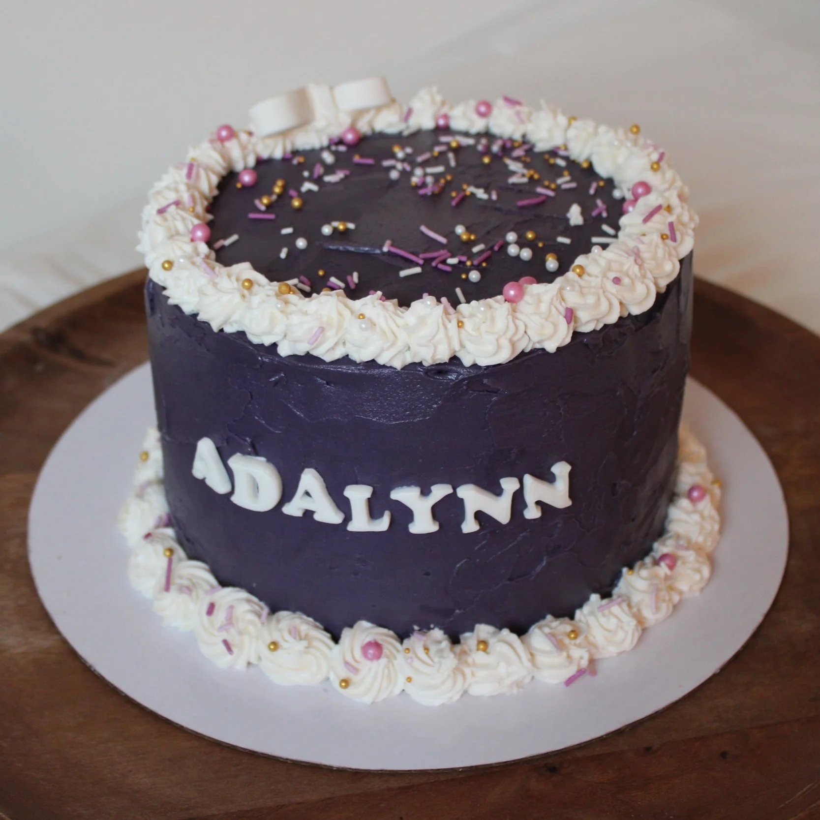 Round cake frosted in deep purple buttercream with white piped borders, pink and gold sprinkles, and the name “Dalynn” displayed in white lettering on the side. The cake is presented on a white cake board atop a wooden serving tray.