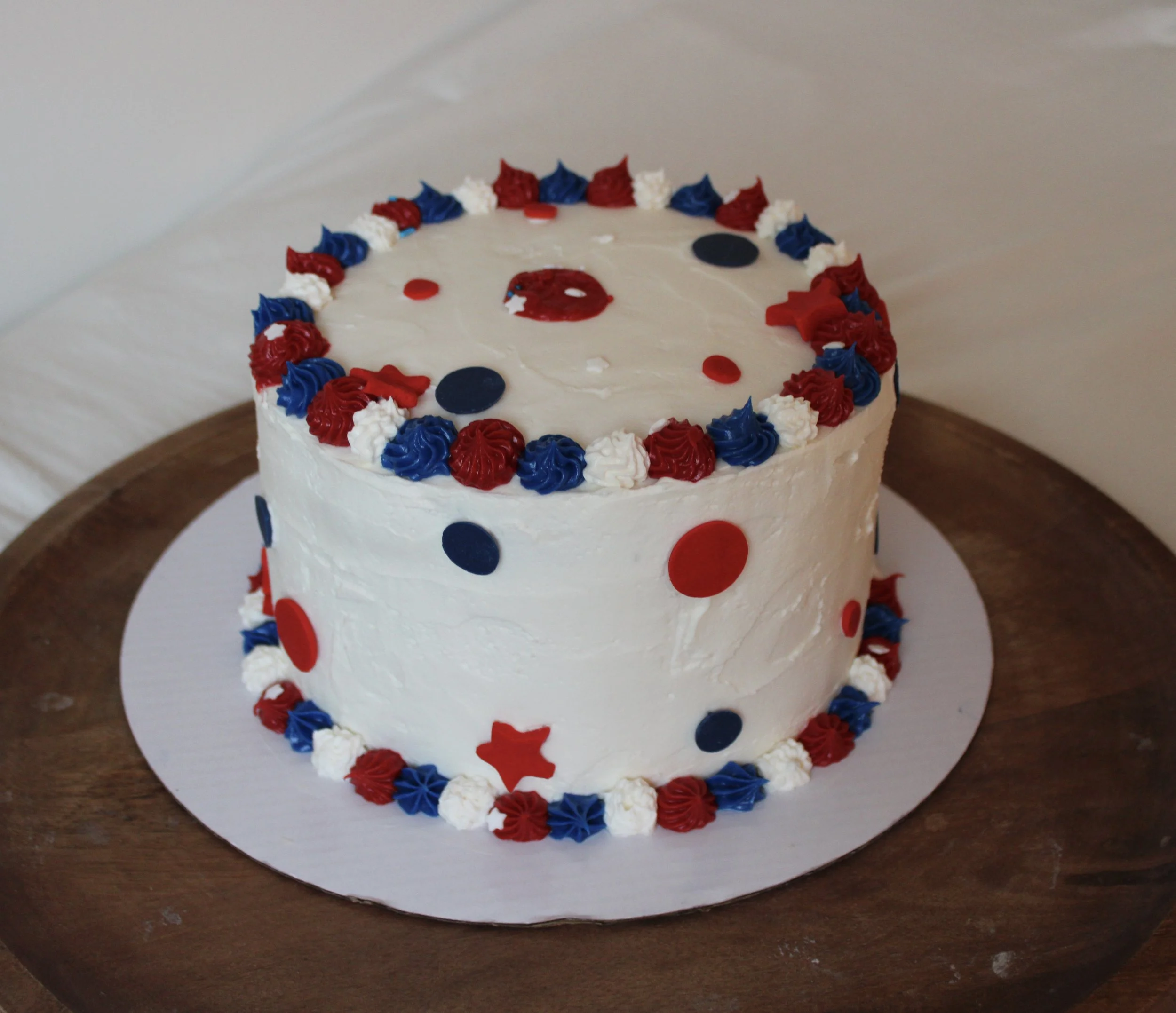 White buttercream cake decorated with red, white, and blue piping, fondant stars and polka dots, and a festive patriotic design. The cake sits on a white cake board atop a wooden serving tray.